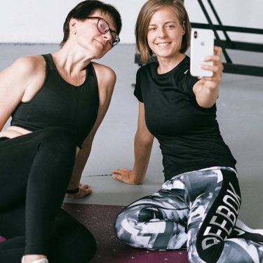 Two women in athletic clothing sitting on a yoga mat indoors, capturing a selfie post workout.
